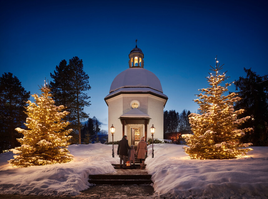 Schneebedeckte Kapelle bei Nacht, flankiert von beleuchteten Weihnachtsbäumen, mit Menschen, die hineingehen.