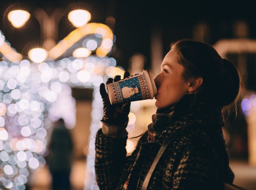 Eine Frau trinkt aus einem Weihnachtspappbecher und genießt die festliche Beleuchtung bei Nacht.