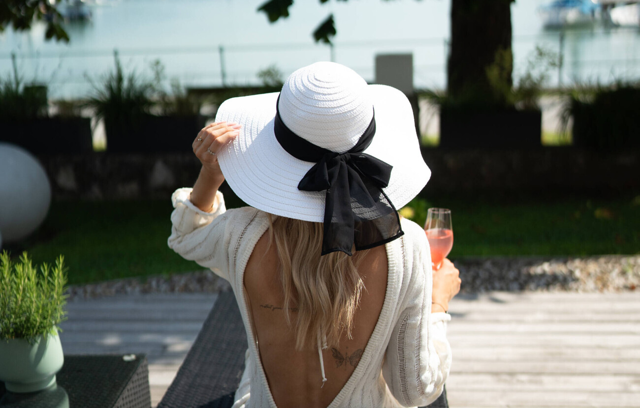 Eine Frau mit Hut sitzt auf der Seeterrasse und hält ein Glas Prosecco in der Hand