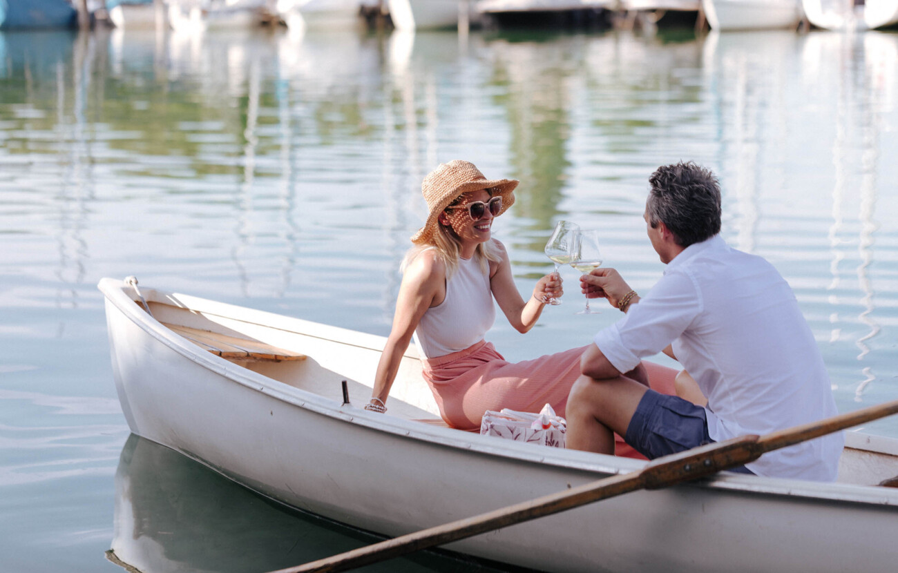 Eine Frau und ein Mann sitzen in einem Ruderboot und stoßen mit einem Glas Sekt an.