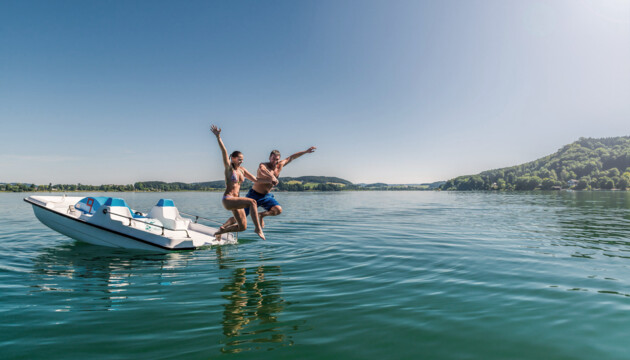 Zwei Personen springen von einem Tretboot ins Wasser des Mattsees bei sonnigem Wetter.