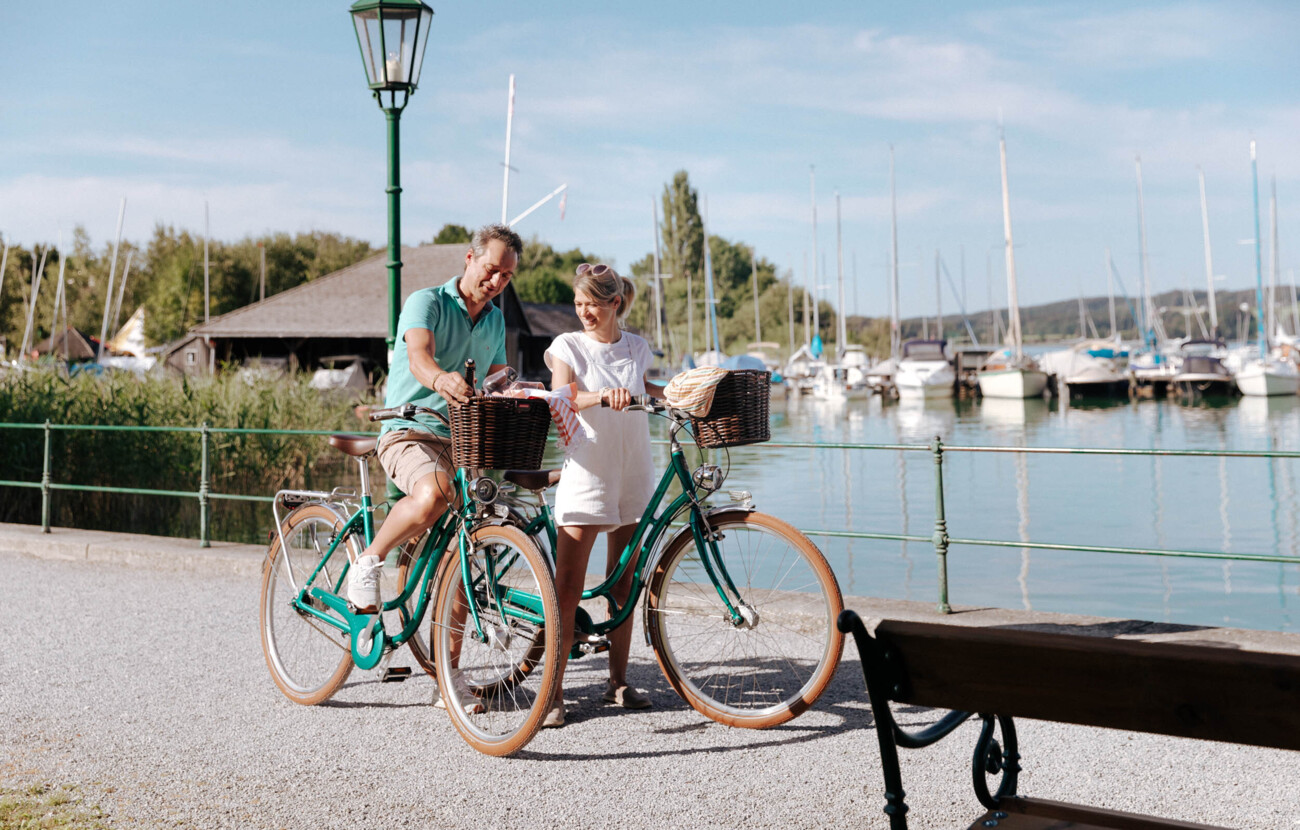 Ein Paar genießt eine Fahrradtour am Ufer eines Sees, im Hintergrund sind Boote zu sehen.