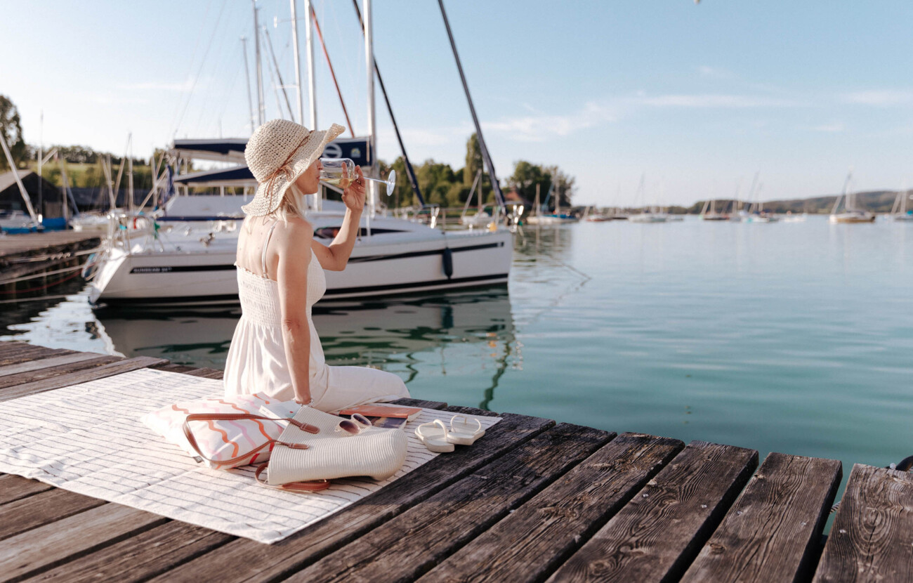 Frau in Sommerkleid und Hut genießt ein Getränk am Steg mit Blick auf Segelboote und den Mattsee.