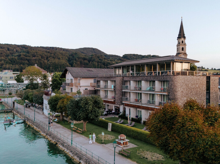 Hotel mit Blick auf den Mattsee, umgeben von Bäumen und einer Uferpromenade. Im Hintergrund ist ein Kirchturm zu sehen.