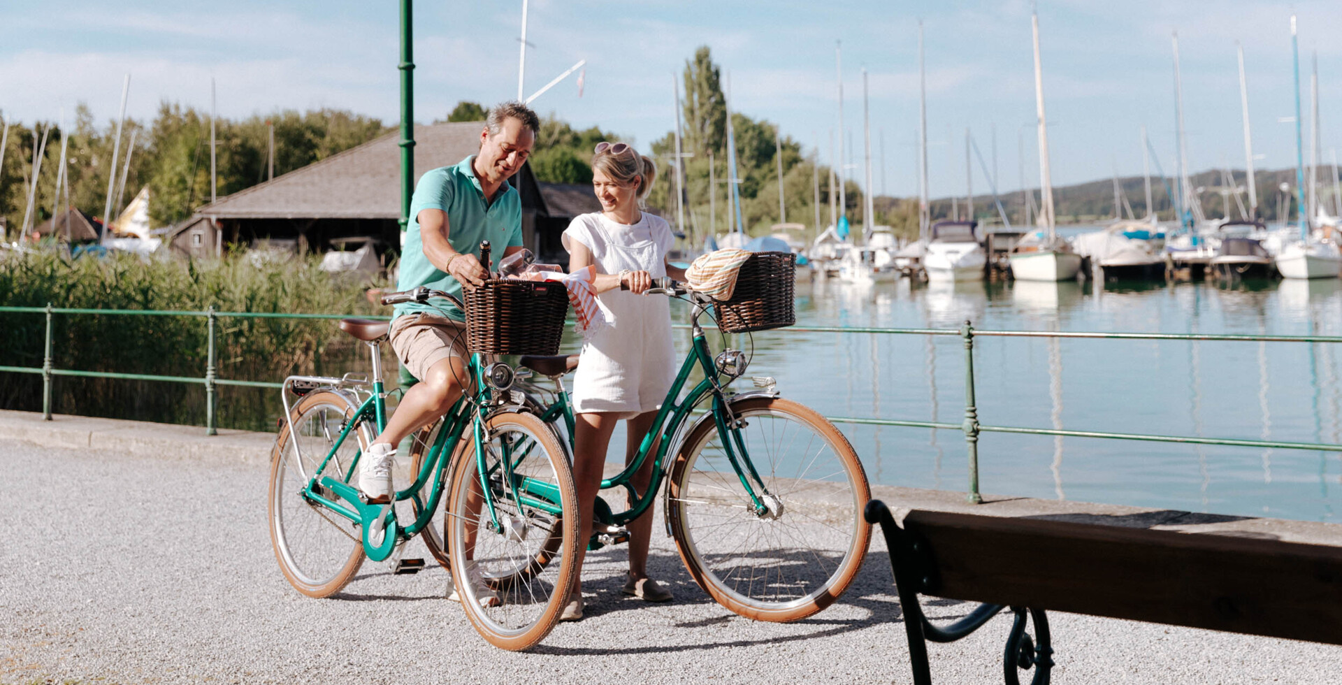 Ein Paar genießt eine Fahrradtour am Ufer eines Sees, im Hintergrund sind Boote zu sehen.