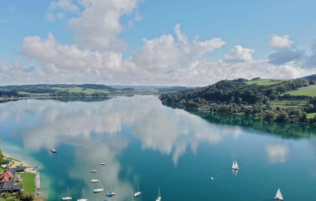 Luftaufnahme des Mattsees mit Segelbooten und bewaldeten Hügeln im Hintergrund, nahe dem Seewirt Mattsee Hotel.