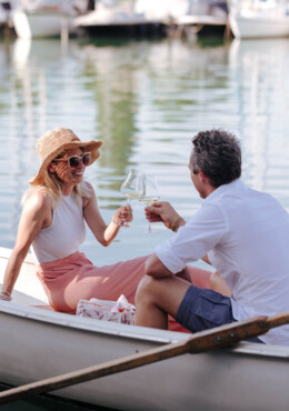 Ein Paar genießt ein Glas Wein in einem Ruderboot auf dem Wasser, umgeben von Segelbooten im Hafen.