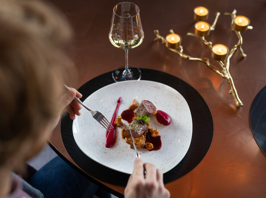 Eine Person genießt ein Gourmetgericht mit Rotweinsoße und einem Glas Weißwein im Seewirt Mattsee. Elegante Tischdekoration mit Kerzen.