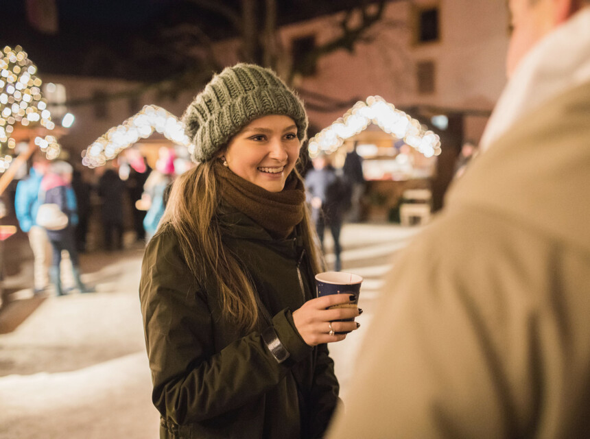 Eine Frau mit Mütze hält ein Getränk auf einem festlich beleuchteten Weihnachtsmarkt.