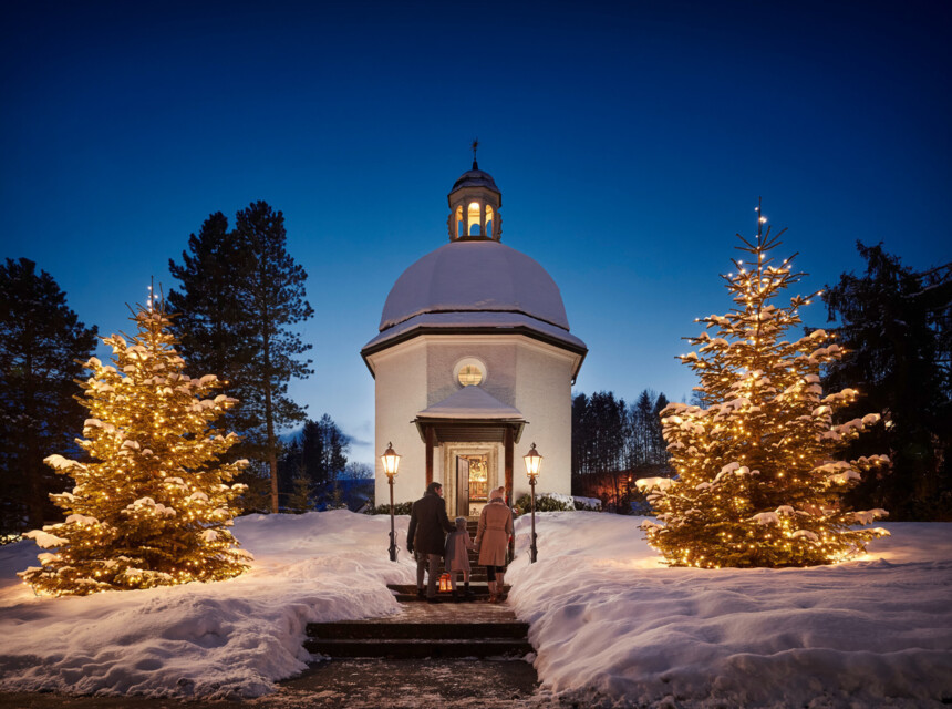 Schneebedeckte Kapelle bei Nacht, flankiert von beleuchteten Weihnachtsbäumen, mit Menschen, die hineingehen.