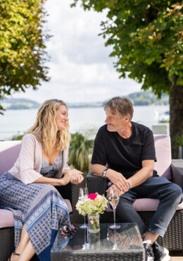 Zwei Personen sitzen auf einem Sofa im Freien am See, genießen Getränke und unterhalten sich. Im Hintergrund sind Bäume und Wasser zu sehen.