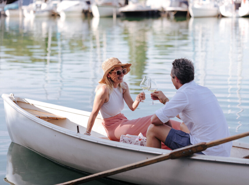 Ein Paar genießt ein Glas Wein in einem Ruderboot auf dem Wasser, umgeben von Segelbooten im Hafen.