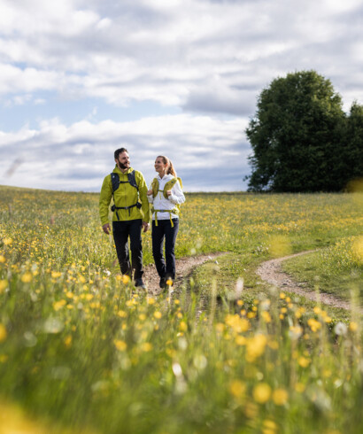 Ein Paar wandert auf einem blumigen Pfad in einer grünen Landschaft unter bewölktem Himmel.