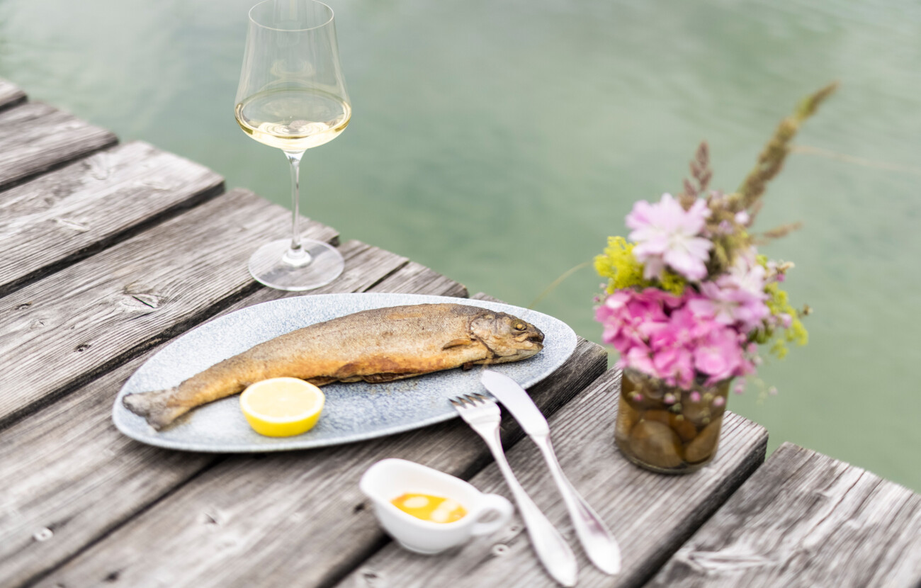 Gegrillter Fisch mit Zitrone, Weißweinglas und Blumenarrangement auf Holztisch am Seeufer von Seewirt Mattsee.