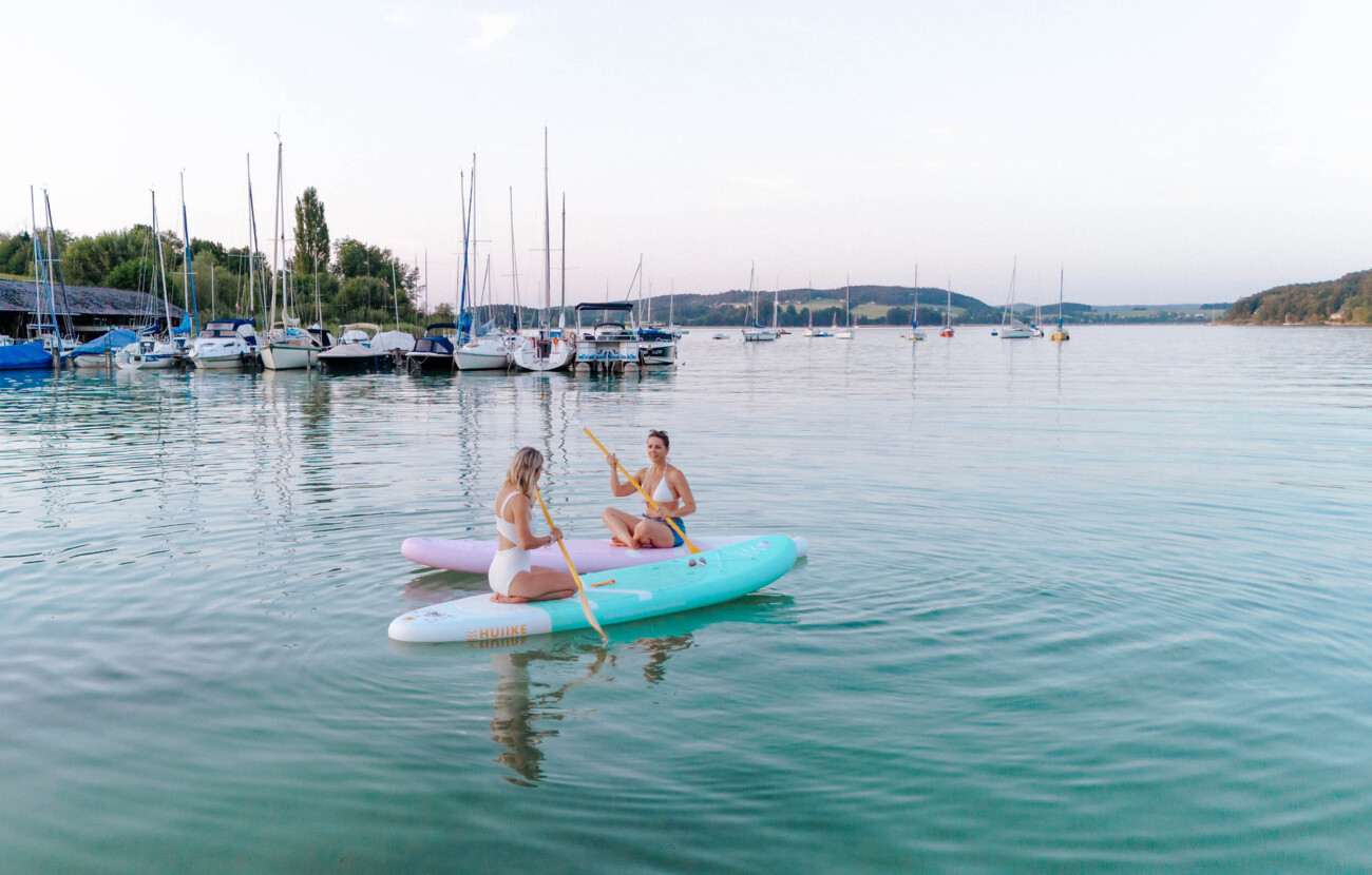 Zwei Personen paddeln auf SUP-Boards auf einem ruhigen See, mit Segelbooten im Hintergrund.