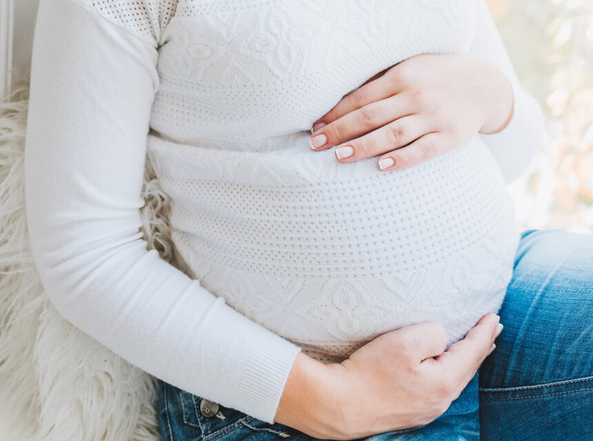 Eine schwangere Frau hält ihren Bauch, bekleidet mit einem weißen Pullover und blauen Jeans, sitzt auf einem weichen Kissen.