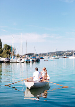 Ein Paar rudert in einem Boot auf einem ruhigen See, umgeben von Segelbooten und blauem Himmel.