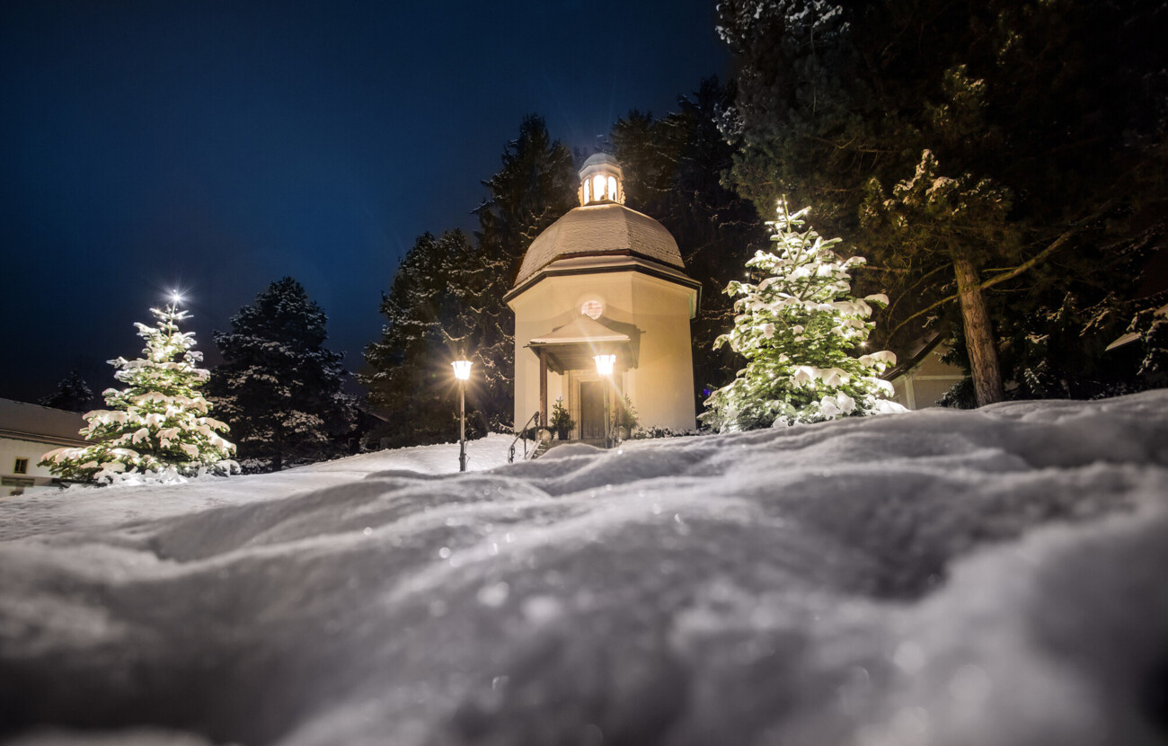 Verschneite Kapelle mit beleuchteten Bäumen bei Nacht in Mattsee.