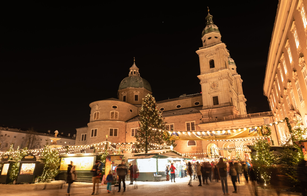 Weihnachtsmarkt bei Nacht vor historischer Kirche, festlich beleuchtet mit Lichterketten und geschmücktem Baum, Menschen schlendern umher.